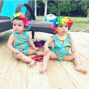 May include: Two babies wearing matching blue and yellow pineapple print swimsuits with rainbow flower headbands. They are sitting on a wooden deck.