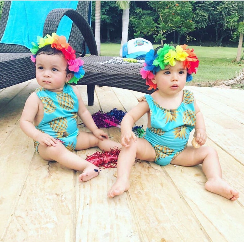 May include: Two babies wearing matching blue and yellow pineapple print swimsuits with rainbow flower headbands. They are sitting on a wooden deck.