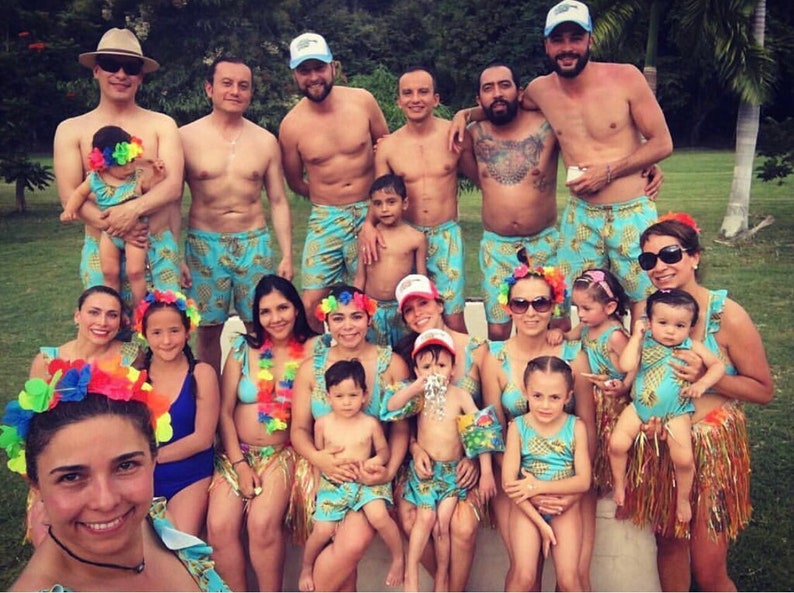 May include: A group of people wearing pineapple-themed swimsuits and accessories. Some are wearing leis, hats, and headbands. There are adults and children in the photo.