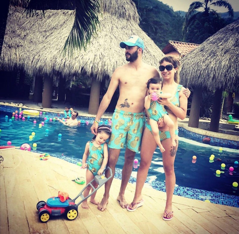 May include: A family of four wearing matching pineapple print swimsuits. The father is wearing a blue baseball cap. The mother is holding a young child in a blue swimsuit. The family is standing by a swimming pool with blue water and colorful floating balls.