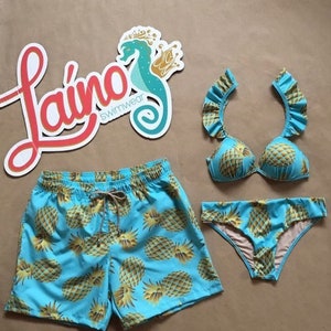 May include: A blue and yellow pineapple print swimsuit with a matching pair of swim trunks. The swimsuit top has ruffled straps and the bottoms are a bikini style. The brand name "La&iacute;no Swimwear" is visible in the image.