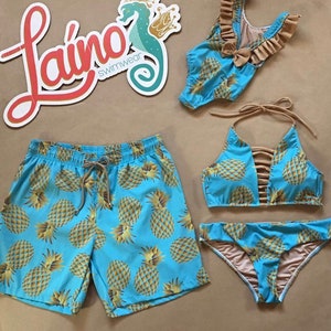 May include: A matching set of swimwear featuring a pineapple print. The set includes a pair of blue swim trunks, a blue bikini top with brown straps, and a blue bikini bottom with brown trim. The logo "Lai&ntilde;o Swimwear" is visible in the background.