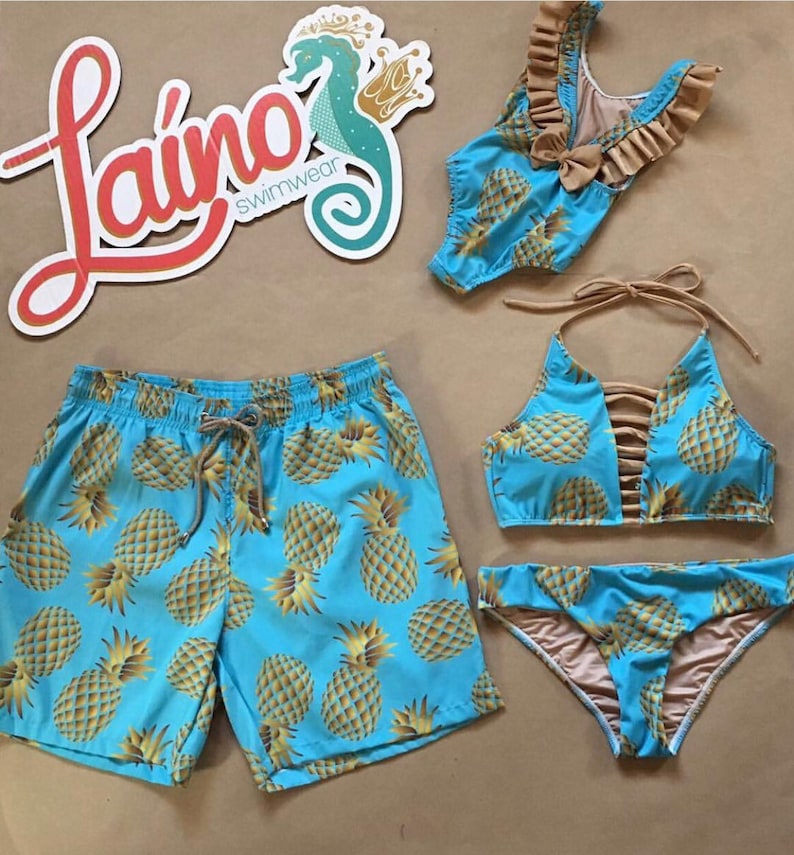 May include: A matching set of swimwear featuring a pineapple print. The set includes a pair of blue swim trunks, a blue bikini top with brown straps, and a blue bikini bottom with brown trim. The logo "Lai&ntilde;o Swimwear" is visible in the background.