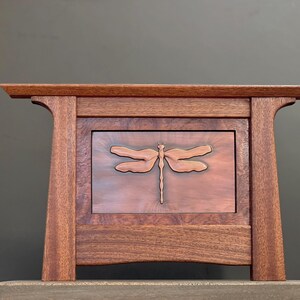May include: A handcrafted wooden box with a dragonfly carving on the front. The box is made of dark brown wood with a copper-colored dragonfly design in the center. The box has a rectangular shape and a flat top.