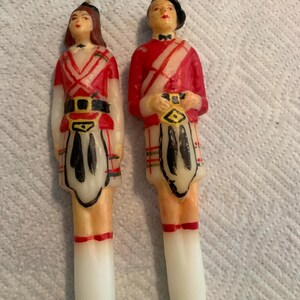 May include: Two vintage wax candles depicting a man and woman in traditional Scottish kilts. The candles are painted in red, white, and black with a plaid pattern.