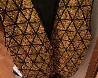 May include: A black vest with gold sequins arranged in a geometric pattern. The vest has a V-neck and is hanging on a hanger. The sequins are sewn onto a black fabric, creating a striking contrast.