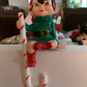 May include: A vintage Christmas elf figurine with a red and white Santa hat, green tunic, and red boots. The elf is holding a white candy cane with red stripes. The figurine has a painted face and is likely made of plastic.