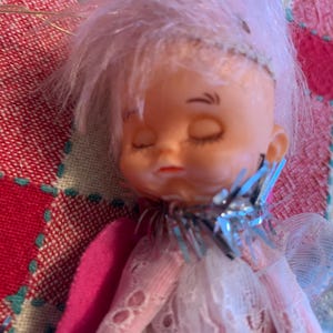 May include: A vintage doll with closed eyes and pink hair. The doll is wearing a white lace dress with a pink felt wing and a silver collar. The doll is a collectible item.