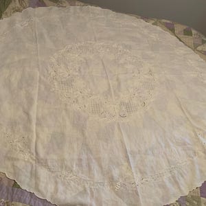 May include: A white round tablecloth with delicate lace embroidery. The tablecloth has a scalloped edge and a central design of floral motifs.