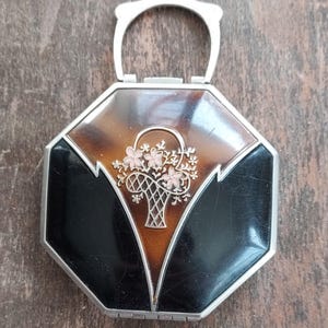 May include: Vintage octagonal compact with a silver handle and trim. The top features a tortoiseshell design with black accents and a floral basket motif in pink and gold.