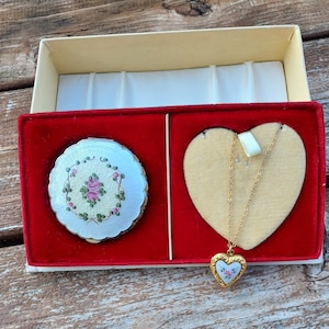 May include: A red velvet box with a white and gold heart-shaped locket and a white and gold compact with a floral design. The locket and compact are both decorated with pink roses.