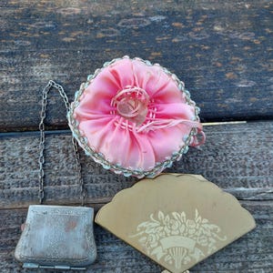 May include: An antique collection of vintage items, including a pink pincushion, a silver chain purse, and a fan-shaped compact. The compact is beige with a floral design. The items are arranged on a weathered wooden surface.