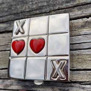 May include: A silver tic-tac-toe game with two red hearts and an X in the bottom right square.