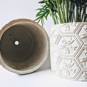 Puede incluir: Dos macetas de cer&aacute;mica blanca, una con superficie lisa y la otra con un patr&oacute;n geom&eacute;trico en relieve de hex&aacute;gonos y flores. Ambas macetas tienen un agujero de drenaje en la parte inferior.