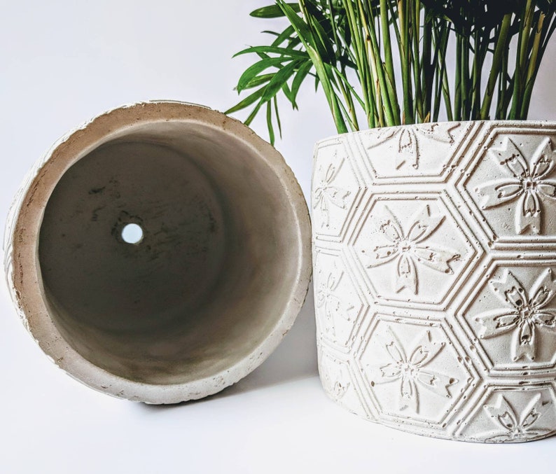 Puede incluir: Dos macetas de cer&aacute;mica blanca, una con superficie lisa y la otra con un patr&oacute;n geom&eacute;trico en relieve de hex&aacute;gonos y flores. Ambas macetas tienen un agujero de drenaje en la parte inferior.