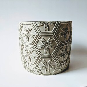Puede incluir: Un macetero de hormig&oacute;n gris claro con un patr&oacute;n hexagonal y dise&ntilde;os de flores en relieve.