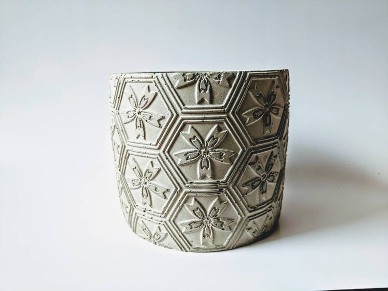 Puede incluir: Un macetero de hormig&oacute;n gris claro con un patr&oacute;n hexagonal y dise&ntilde;os de flores en relieve.