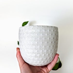 Op de afbeelding: Een witte keramische plantenpot met een geometrisch patroon. De pot heeft de vorm van een cilinder en heeft een gladde, matte afwerking.