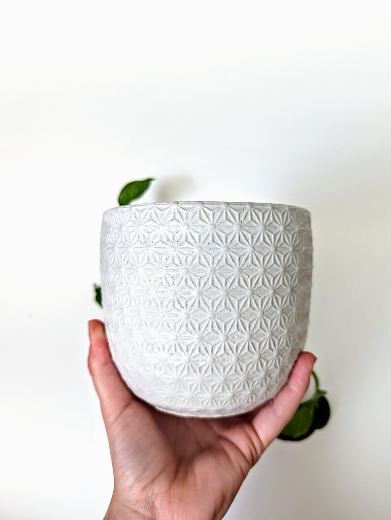 Op de afbeelding: Een witte keramische plantenpot met een geometrisch patroon. De pot heeft de vorm van een cilinder en heeft een gladde, matte afwerking.