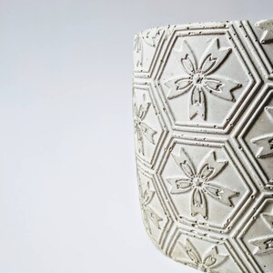 Puede incluir: Maceta de cer&aacute;mica blanca con un patr&oacute;n hexagonal y dise&ntilde;os de flores en relieve.