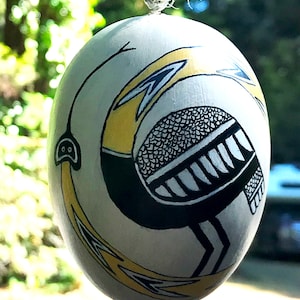 May include: A white egg-shaped ornament with a black and yellow bird design. The bird has a long neck and a long beak. The bird is surrounded by yellow and black lines.
