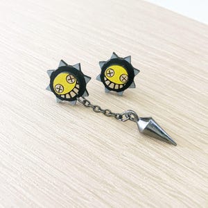 May include: A pair of unique earrings featuring a yellow face with a stitched smile and cross-shaped eyes, surrounded by a black spiky border. One earring is connected to a silver spike charm by a chain.