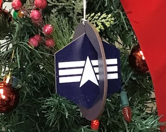 May include: A blue and white Christmas bauble shaped like a shield with a star and three stripes. The bauble is hanging on a green Christmas tree.