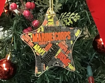 May include: A star-shaped Christmas bauble with a gold string for hanging. The bauble is made of clear plastic and features a word cloud design with the words "Marine Corps" in large, bold letters. Other words in the design include "Adapt", "Improvise", "Semper Fi", and "HooRah!"