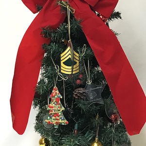 Puede incluir: Un pequeño árbol de Navidad artificial decorado con adornos, incluyendo un lazo rojo, una estrella dorada, una piña y una bola transparente con un árbol de Navidad dentro. El árbol también está decorado con una insignia de rango militar amarilla y verde.