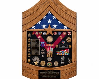 May include: A wooden display case shaped like a Sergeant Major rank insignia, featuring a folded American flag, various military medals, pins, ribbons, and dog tags on a black background. A plaque at the bottom reads SGM Stern Loe Lex.