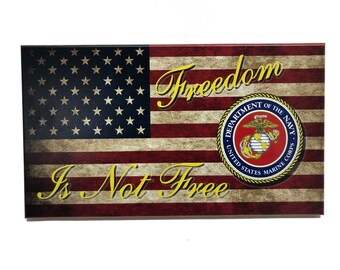 May include: A distressed American flag with the words "Freedom Is Not Free" in gold lettering. The flag has a blue circle with the United States Marine Corps emblem in the centre.