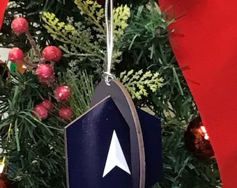 May include: A blue and white wooden Christmas bauble shaped like a hexagon with a white arrow pointing upwards. The bauble is hanging on a Christmas tree.