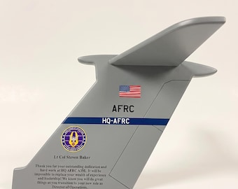 May include: A grey model of an aircraft tail fin, with the American flag and the text "AFRC HQ-AFRC." The model includes a dedication to Lt Col Steven Baker, with a message about his service and new role as Director of Operations.
