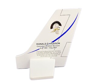 May include: A white wooden aeroplane tail fin with a blue stripe and a gold helmet graphic. The text "RONALD D. HUDSON Centurion Aviation Services Aug 1984 - Aug 2021" is printed on the fin.