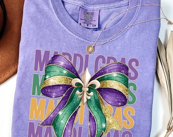 May include: A purple, green and gold Mardi Gras themed t-shirt featuring a large bow and fleur-de-lis design. The shirt has the words "Mardi Gras" repeated in a stacked pattern.