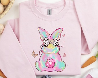 May include: Pink sweatshirt with a pastel rainbow bunny wearing a leopard print bow. The bunny has a pink flower on its chest. The sweatshirt is on a white surface with flowers and Easter eggs.