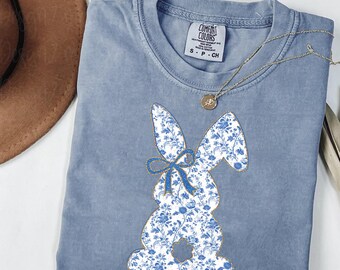 May include: A light blue t-shirt with a floral patterned bunny silhouette. The bunny has a blue bow and is centered on the shirt. The shirt has a tag that reads "Comfort Colors" and "S-P-CH".
