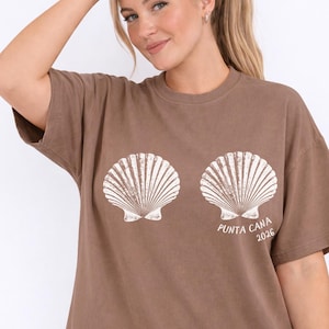 May include: A brown t-shirt featuring two white seashell graphics and the text "PUNTA CANA 2026". The shirt has short sleeves and a relaxed fit, suitable for casual wear.