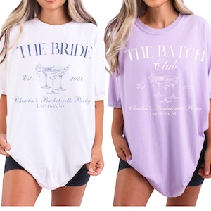May include: Two oversized t-shirts, one white and one lavender, with text and graphics. The white shirt reads "THE BRIDE" above a cocktail graphic and "Claudia's Bachelorette Party LAS VEGAS, NV." The lavender shirt says "THE BATCH Club" with similar design elements.