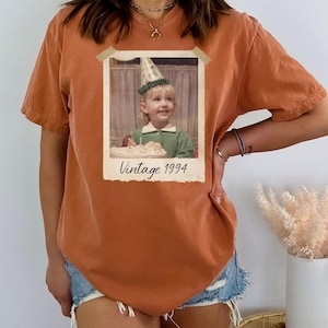 Puede incluir: Camiseta de color naranja quemado con estampado de foto vintage. La foto muestra a un ni&ntilde;o peque&ntilde;o con un gorro de fiesta, con el texto "Vintage 1994" debajo. La camiseta tiene mangas cortas y un corte holgado.