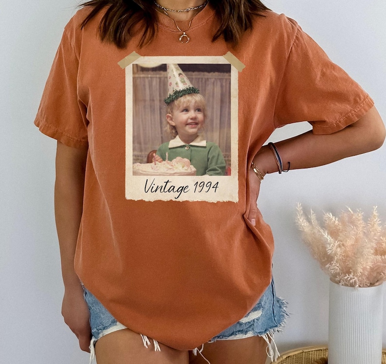 Puede incluir: Camiseta de color naranja quemado con estampado de foto vintage. La foto muestra a un ni&ntilde;o peque&ntilde;o con un gorro de fiesta, con el texto "Vintage 1994" debajo. La camiseta tiene mangas cortas y un corte holgado.