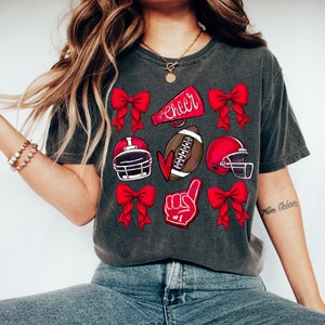 May include: Dark gray t-shirt featuring red bows, football helmets, a football, a foam finger with "#1", and a megaphone with the word "Cheer". The shirt is a comfortable, casual style.