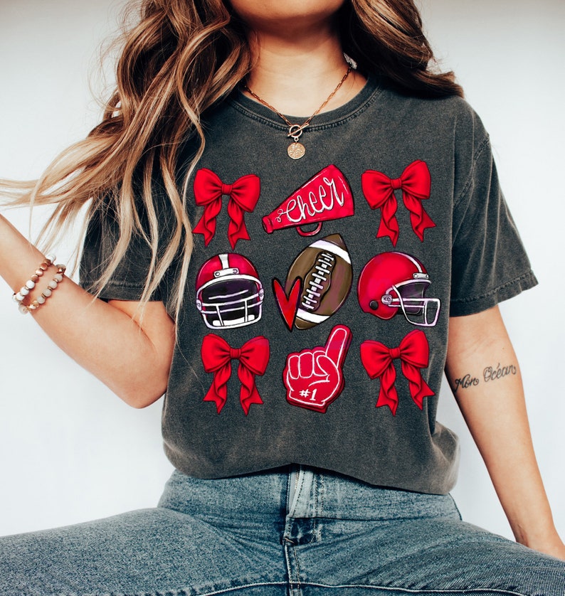 May include: Dark gray t-shirt featuring red bows, football helmets, a football, a foam finger with "#1", and a megaphone with the word "Cheer". The shirt is a comfortable, casual style.
