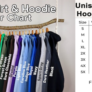May include: A color chart for sweatshirts and hoodies, displaying a variety of colors hanging on a wooden branch. The chart includes colors like white, pink, red, green, and blue. A size chart is also included, with measurements in inches for width, length, and sleeve.