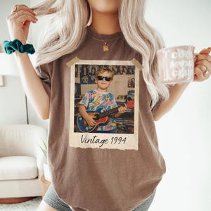 Puede incluir: Una camiseta marr&oacute;n con una impresi&oacute;n fotogr&aacute;fica vintage. La foto muestra a una persona joven con gafas de sol sosteniendo una guitarra. La foto est&aacute; etiquetada como "Vintage 1994". Una taza rosa con el texto "Cool Mom Club" se sostiene en la mano.