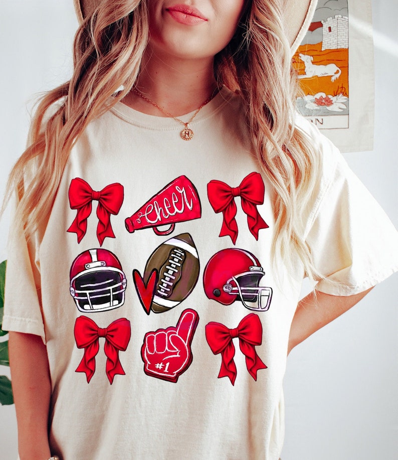 May include: Cream-colored t-shirt featuring a collection of red bows, football helmets, a football, a foam finger, and a megaphone with the word "Cheer". The shirt is a comfortable, casual style.