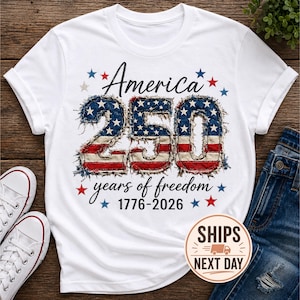 May include: White t-shirt with the text "America 250 years of freedom 1776-2026" in black script. The number "250" is designed with a distressed American flag pattern. Red and blue stars surround the text.