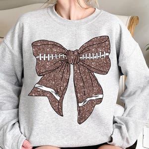 Può includere: Una felpa grigio chiaro con un grande fiocco marrone glitterato. Il fiocco è disegnato per assomigliare a un pallone da football, completo di cuciture e strisce bianche. Il disegno è centrato sulla parte anteriore della felpa.