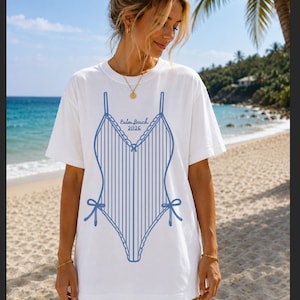 May include: White t-shirt featuring a blue swimsuit design with vertical stripes, bows, and the text "Palm Beach 2026." The shirt is worn on a beach with a blue ocean and palm trees in the background.