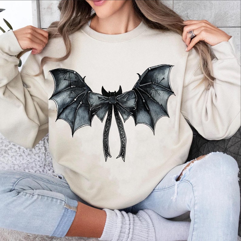 May include: A cream-coloured sweatshirt with a black bat wing and bow design. The bat wings have detailed shading and a textured appearance. The bow has long, flowing ribbons. A unique and stylish top for Halloween or gothic-themed outfits.